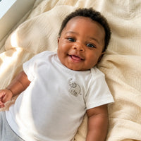 Bébé sénégalais souriant portant un t-shirt en coton blanc à manches courtes, allongé sur une couverture douce dans une maison de Dakar
